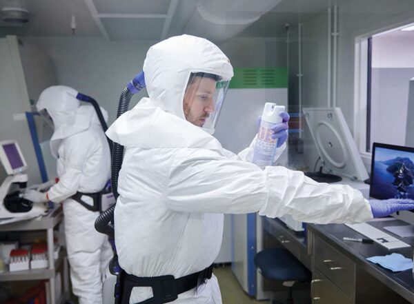 Two postdoctoral researchers wearing white biohazard suits and purple gloves work on the novel coronavirus in a biosafety level-3 lab. In the foreground, one of the researchers holds out his arm to spray his suit with an aerosol from a can.