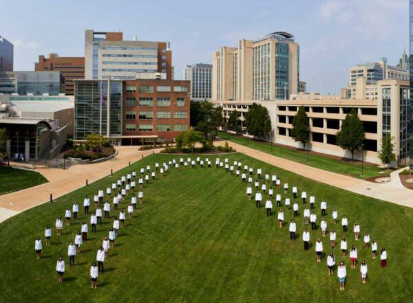 First-year medical students pose for a socially distanced class portrait in their new white coats. The students are lined up on a green lawn in the shape of an arch, to represent the rollout of the new Gateway Curriculum.