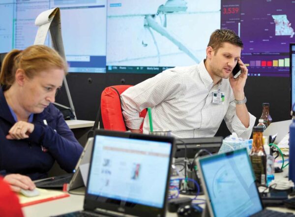 Team members on their phone and at the computers, keep their calm surrounded by large screens, more computers, and bottles of water and caffeine