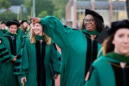 A young Black woman in emerald green regalia smiles broadly and points to an acquaintance as she walks in the Commencement processional